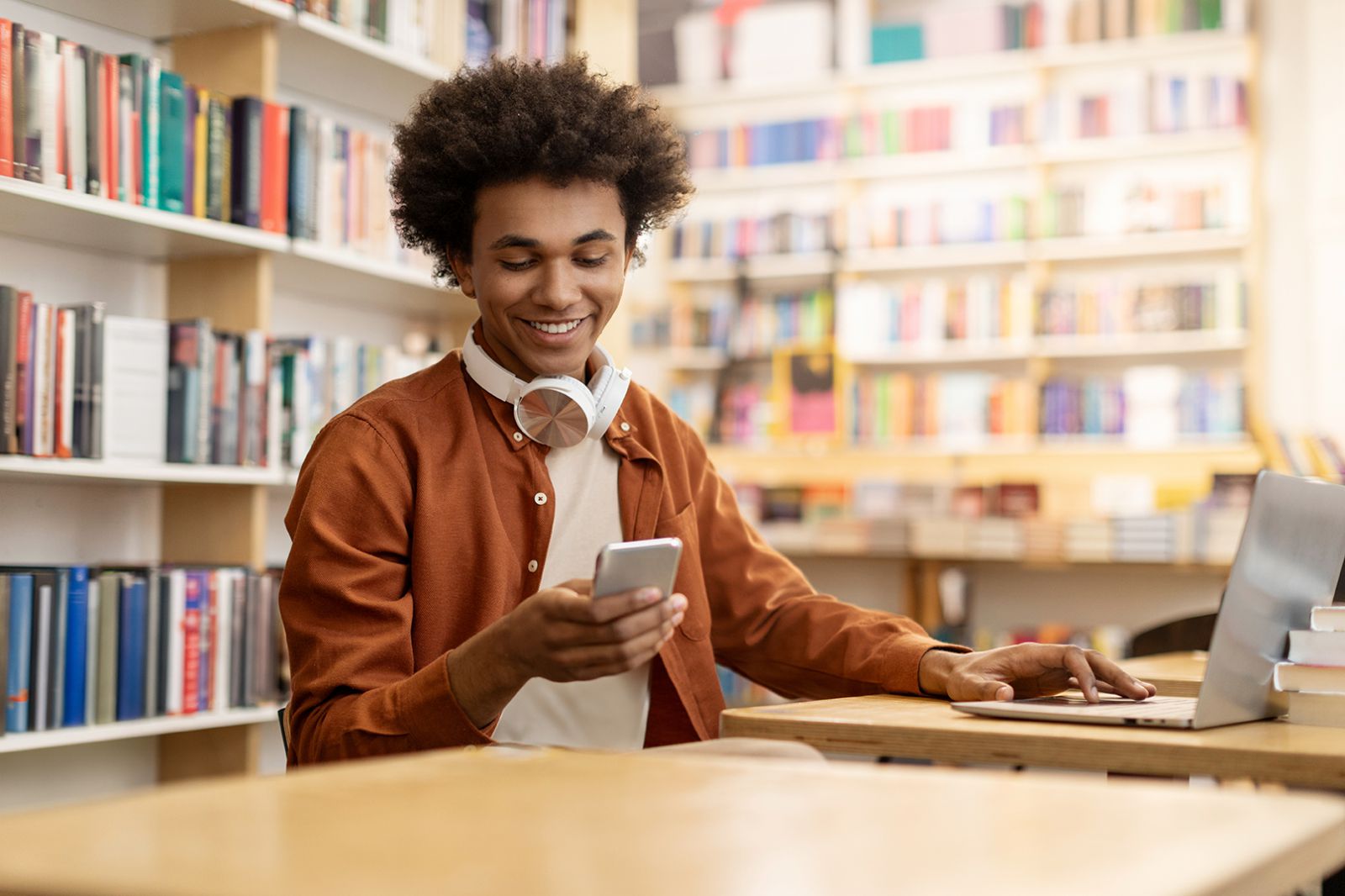 A young adult student in a library using a laptop smiles as he looks at a cellphone.